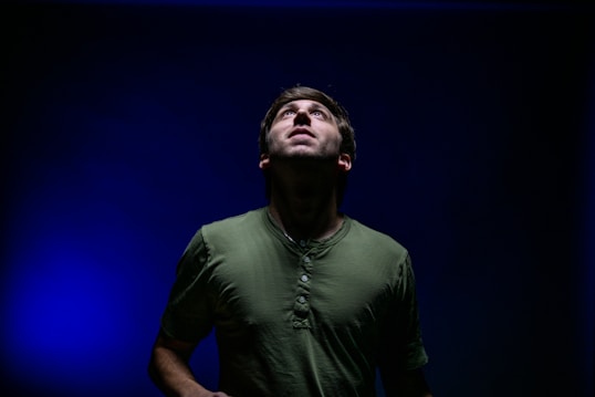A person in a green shirt gazes upwards with an expression of contemplation or wonder. The background is dark, with a vibrant blue light creating a dramatic and contrasting effect.