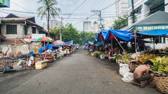 A bustling street market scene with various vendors on both sides displaying fruits and vegetables under makeshift blue and red tarpaulins. People are sitting and engaging in activities at different stalls. A palm tree and urban buildings are visible in the background, reflecting a mix of urban and street life.