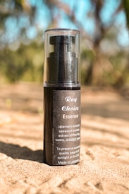 A small black bottle of 'Ray Choice Essence' is placed on sandy ground with a blurred background of nature, including possibly trees and foliage. The label on the bottle mentions various extracts like jatamansi, oakwood, and coffee beans, and advises on how to preserve the essence quality.