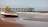 A jet ski is parked on a sandy beach near the water's edge. In the background, a pier extends into the ocean with several people standing on it. The sky is clear with a few clouds and the ocean waves are gently crashing onto the shore.