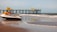 A jet ski is parked on a sandy beach near the water's edge. In the background, a pier extends into the ocean with several people standing on it. The sky is clear with a few clouds and the ocean waves are gently crashing onto the shore.