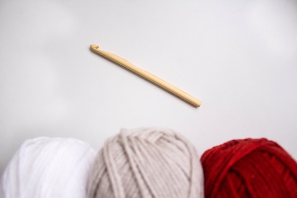 Three skeins of yarn in white, beige, and red are positioned at the bottom of the image, with a wooden crochet hook lying diagonally above them on a light gray background.