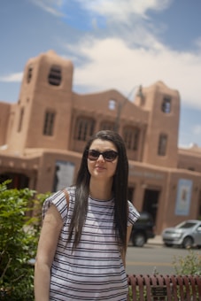 A woman with long dark hair wearing sunglasses and a striped shirt stands in front of an adobe-style building. The sky is partly cloudy, and there is vegetation on the left side. The building has a distinctive southwestern architectural style with earthy tones.