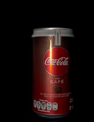 A can of Coca-Cola with Coffee is displayed against a black background. The can has a red and brown design with the Coca-Cola logo prominently featured and text indicating it's an espresso flavor. The nutritional information is visible at the bottom.