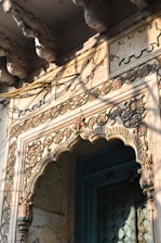 An intricately carved stone archway features floral and geometric patterns with a mix of weathered colors. The structure displays detailed craftsmanship, possibly from a historic or cultural building. Light and shadows play across the carvings, highlighting their depth and texture.