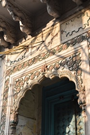 An intricately carved stone archway features floral and geometric patterns with a mix of weathered colors. The structure displays detailed craftsmanship, possibly from a historic or cultural building. Light and shadows play across the carvings, highlighting their depth and texture.