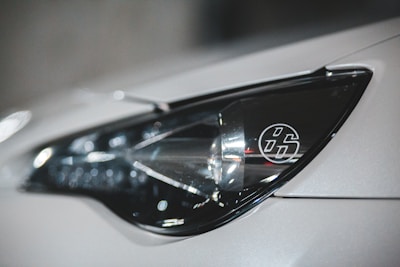 Close-up view of a car's headlight with a distinctive emblem featuring the number 86 inside a circle. The headlight design is modern and sleek, with a reflective surface capturing light reflections.
