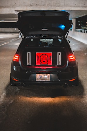 A black car is parked with the trunk open in a dimly lit parking garage. Inside the trunk, a red backlit panel displays a graphic with the words 'ALERT MAFIA'. The car's rear lights illuminate the surrounding area, and the license plate is visible. Concrete walls surround the parking area with some night ambient light shining in from the side.