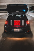 A black car is parked with the trunk open in a dimly lit parking garage. Inside the trunk, a red backlit panel displays a graphic with the words 'ALERT MAFIA'. The car's rear lights illuminate the surrounding area, and the license plate is visible. Concrete walls surround the parking area with some night ambient light shining in from the side.