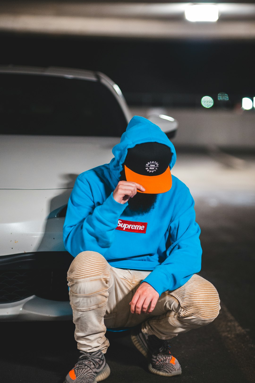 man in blue hoodie wearing orange and black cap sitting on white car during daytime