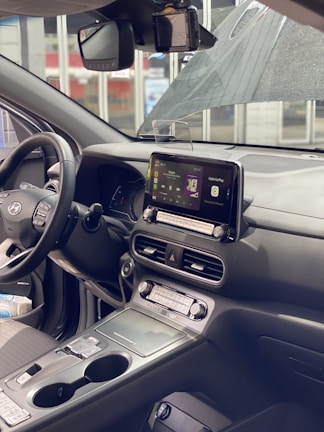 The interior of a modern car featuring a digital dashboard, infotainment system with Apple CarPlay displayed, and a steering wheel. The dashboard includes various buttons and dials, and there is a large sunroof visible overhead. The seats appear to be cloth upholstered, and there are items stored in the console.