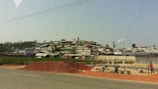 A settlement of makeshift structures with corrugated roofs stretches across a hillside. In the foreground, a pile of red dirt is on the side of a road, with workers busy at a construction site. The landscape is a mix of green vegetation and scattered trees.