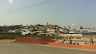A settlement of makeshift structures with corrugated roofs stretches across a hillside. In the foreground, a pile of red dirt is on the side of a road, with workers busy at a construction site. The landscape is a mix of green vegetation and scattered trees.