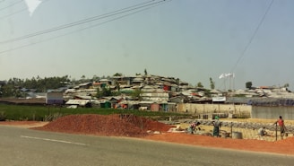 A settlement of makeshift structures with corrugated roofs stretches across a hillside. In the foreground, a pile of red dirt is on the side of a road, with workers busy at a construction site. The landscape is a mix of green vegetation and scattered trees.