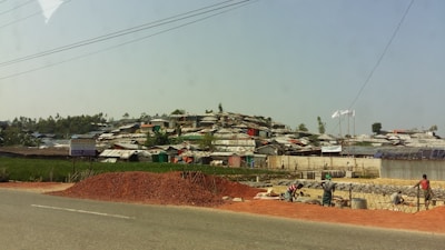 A settlement of makeshift structures with corrugated roofs stretches across a hillside. In the foreground, a pile of red dirt is on the side of a road, with workers busy at a construction site. The landscape is a mix of green vegetation and scattered trees.