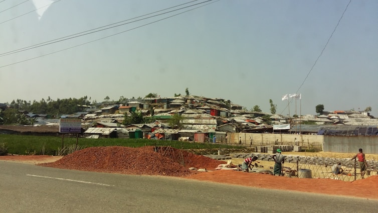 A settlement of makeshift structures with corrugated roofs stretches across a hillside. In the foreground, a pile of red dirt is on the side of a road, with workers busy at a construction site. The landscape is a mix of green vegetation and scattered trees.