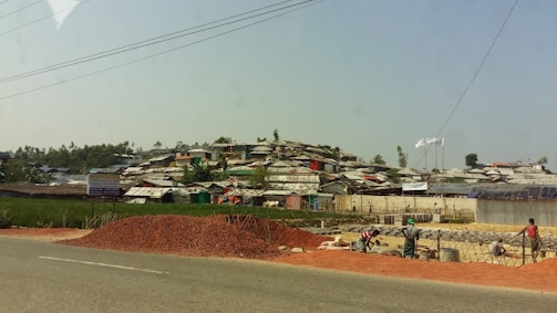 A settlement of makeshift structures with corrugated roofs stretches across a hillside. In the foreground, a pile of red dirt is on the side of a road, with workers busy at a construction site. The landscape is a mix of green vegetation and scattered trees.