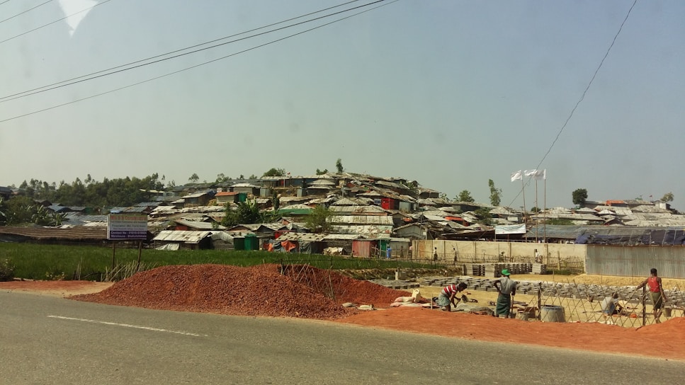 A settlement of makeshift structures with corrugated roofs stretches across a hillside. In the foreground, a pile of red dirt is on the side of a road, with workers busy at a construction site. The landscape is a mix of green vegetation and scattered trees.