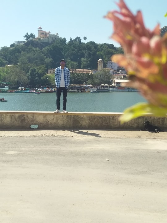 man in blue denim jeans standing on gray concrete pavement near body of water during daytime in Mount Abu India
