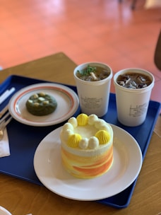 On a blue tray, there is a round cake with yellow and white swirls topped with small dollops of cream and decorative swirls. Next to it, a cup of iced coffee and a bowl of soup are placed, both in white paper cups. A small green pastry is also visible on a white plate with an orange rim.