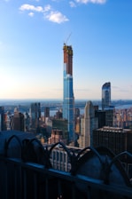 A cityscape featuring a prominent skyscraper under construction, with a crane at the top. The skyline is filled with various high-rise buildings, and the scene is set against a bright blue sky with a few clouds. In the foreground, there is an ornamental railing, suggesting the photo was taken from a high vantage point.