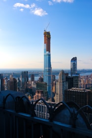 A cityscape featuring a prominent skyscraper under construction, with a crane at the top. The skyline is filled with various high-rise buildings, and the scene is set against a bright blue sky with a few clouds. In the foreground, there is an ornamental railing, suggesting the photo was taken from a high vantage point.
