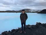 A person stands near a geothermal spa with milky blue water surrounded by dark volcanic rocks. In the background, there are low-lying hills and a cloudy sky. Industrial structures can be seen emitting steam, suggesting geothermal activity.