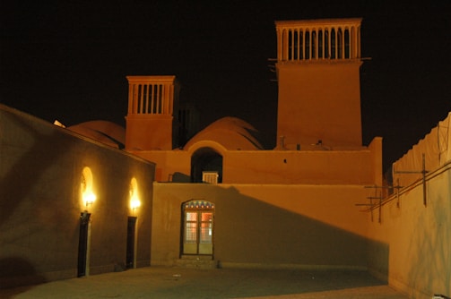 An architectural structure featuring traditional wind towers stands illuminated at night. Warm lights cast soft shadows against beige walls, and decorative windows are subtly visible. The scene is quiet and serene, with an emphasis on cultural and historical architecture.