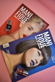 Two magazines are placed on a pink surface. Each magazine features a close-up portrait of a woman with distinct makeup. One magazine cover is predominantly red with a blonde woman wearing red lipstick. The other cover is blue, showcasing a woman with a bob haircut, wearing blue lipstick. There are two bottles of nail polish, one black and one red, placed beside each magazine.