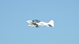 A small white aircraft with the letters 'VCA' on its side is flying mid-air against a clear blue sky. The aircraft has two wings and a propeller that is in motion.