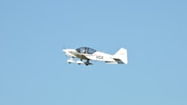 A small white aircraft with the letters 'VCA' on its side is flying mid-air against a clear blue sky. The aircraft has two wings and a propeller that is in motion.