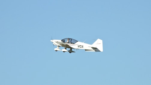 A small white aircraft with the letters 'VCA' on its side is flying mid-air against a clear blue sky. The aircraft has two wings and a propeller that is in motion.