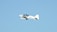 A small white aircraft with the letters 'VCA' on its side is flying mid-air against a clear blue sky. The aircraft has two wings and a propeller that is in motion.