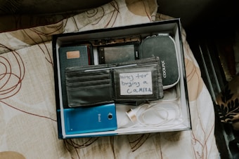 A box contains various items including wallets, a blue mobile device, a digital camera pouch, and a note that says 'money for buying a camera.' The box is placed on a patterned bedspread.