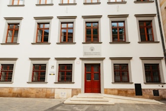 A symmetrical facade of a European academic building featuring multiple windows with red frames. The entrance has a double door, also painted red. The sign above the door indicates it is a faculty of a university. The building is constructed with smooth white walls and trimmed with stone accents. Stone steps lead to the entrance.
