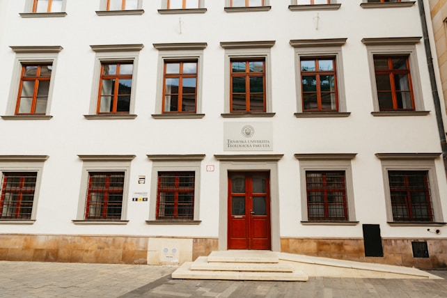 A symmetrical facade of a European academic building featuring multiple windows with red frames. The entrance has a double door, also painted red. The sign above the door indicates it is a faculty of a university. The building is constructed with smooth white walls and trimmed with stone accents. Stone steps lead to the entrance.