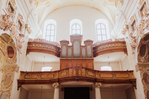 An ornate interior featuring a large pipe organ set against a backdrop of high ceilings and arched windows. Decorative carvings and golden accents adorn the architecture, with intricate sculptures and details visible on the walls. The lighting is soft and natural, entering through the tall windows.