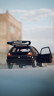 A black Volkswagen Golf GTI with an open driver side door is parked on a concrete surface. The vehicle has a large rear spoiler, and the background features a partially blurred, tall building with windows under a partly cloudy sky.