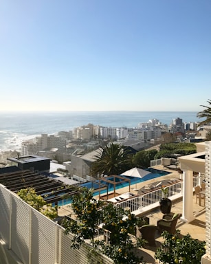 A coastal cityscape with modern architecture featuring numerous buildings near the ocean. In the foreground, there is a luxurious terrace with a pool, outdoor seating, and lush greenery, providing an impressive view of the sea. The horizon shows clear skies with the sun casting a warm glow over the scene.