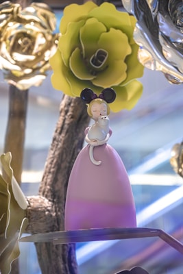 A whimsical figurine of a girl with large black ears, styled in a pink dress, is cradling a small white cat. The backdrop includes oversized flowers and branches, with a prominent green flower above her. The surroundings convey a fantastical, fairy-tale setting.