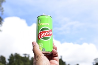 A hand is holding a bright green can labeled 'Limca' with lime and lemon illustrations against a backdrop of a blue sky with scattered white clouds.