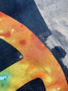A colorful towel or fabric lies partially on sand, featuring a pattern with shades of orange, yellow, and black. The design includes a large, curved shape, possibly part of a letter or symbol. Sand covers a portion of the fabric and the surrounding ground.