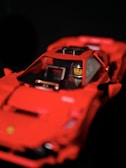 A red toy car resembling a sports car with a figure inside, featuring a yellow face and a helmet. The toy has intricate details, including side mirrors and an interior dashboard.