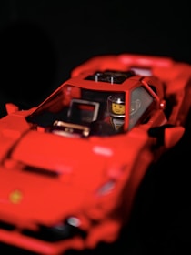 A red toy car resembling a sports car with a figure inside, featuring a yellow face and a helmet. The toy has intricate details, including side mirrors and an interior dashboard.