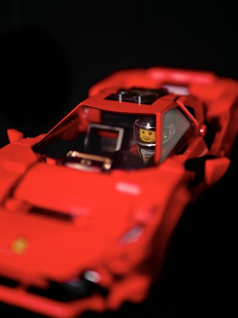 A red toy car resembling a sports car with a figure inside, featuring a yellow face and a helmet. The toy has intricate details, including side mirrors and an interior dashboard.