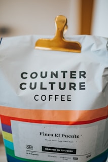 A bag of Counter Culture Coffee clipped with a golden clip, displaying a colorful stripe pattern near the bottom. The label reads 'Finca El Puente' with flavor notes of almond, brown sugar, and baked apple, and indicates a roast date of 7/14/2020.