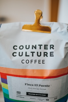 A bag of Counter Culture Coffee clipped with a golden clip, displaying a colorful stripe pattern near the bottom. The label reads 'Finca El Puente' with flavor notes of almond, brown sugar, and baked apple, and indicates a roast date of 7/14/2020.