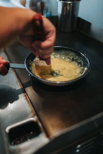A person stirring a creamy mixture in a frying pan on a stovetop using a spatula. The hand holding the spatula is visibly active and focused, with the stovetop and pan dominating the scene.