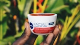 A hand holding a container of NIVEA facial cream against a vibrant green background, likely foliage. The product label is visible, displaying its benefits and contents.