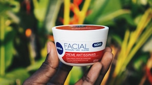 Hands holding a small jar of nourishing cream, surrounded by green leaves and natural textures.